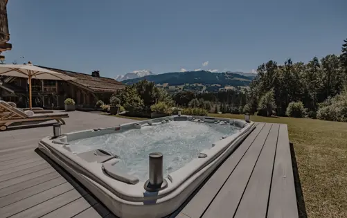 Luxury chalet terrace with outdoor jacuzzi, sun loungers, and panoramic mountain view under a clear blue summer sky.