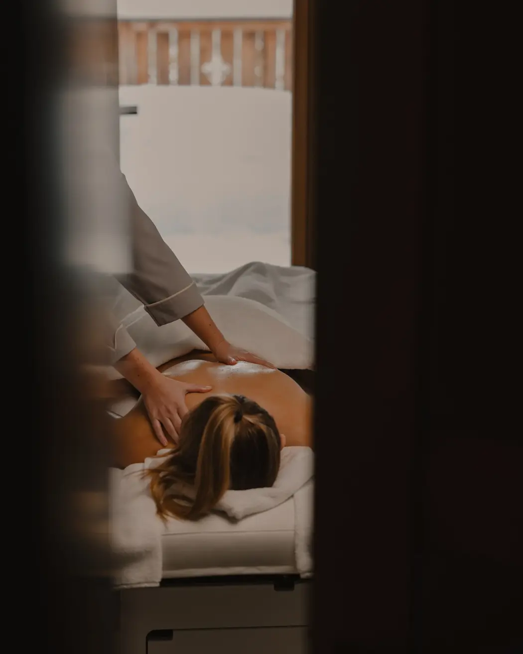 Person receiving a relaxing back massage indoors, with soft lighting and a view of a snowy wooden balcony outside.