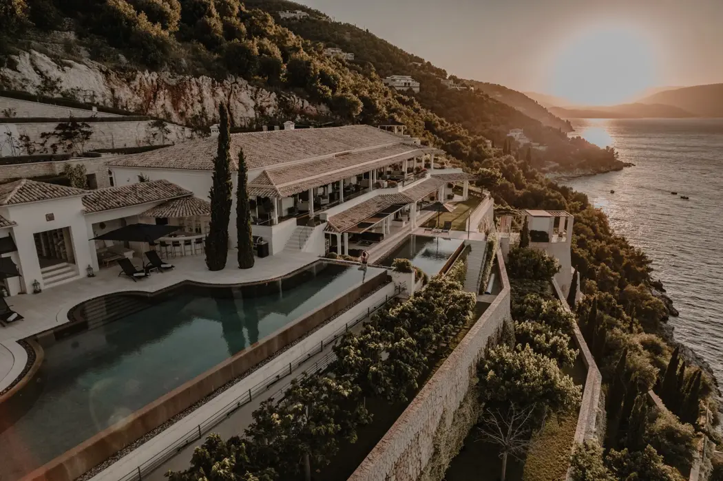 Aerial view of a luxury seaside villa built on a hillside, with terraces, pools, and a sunset over the ocean.