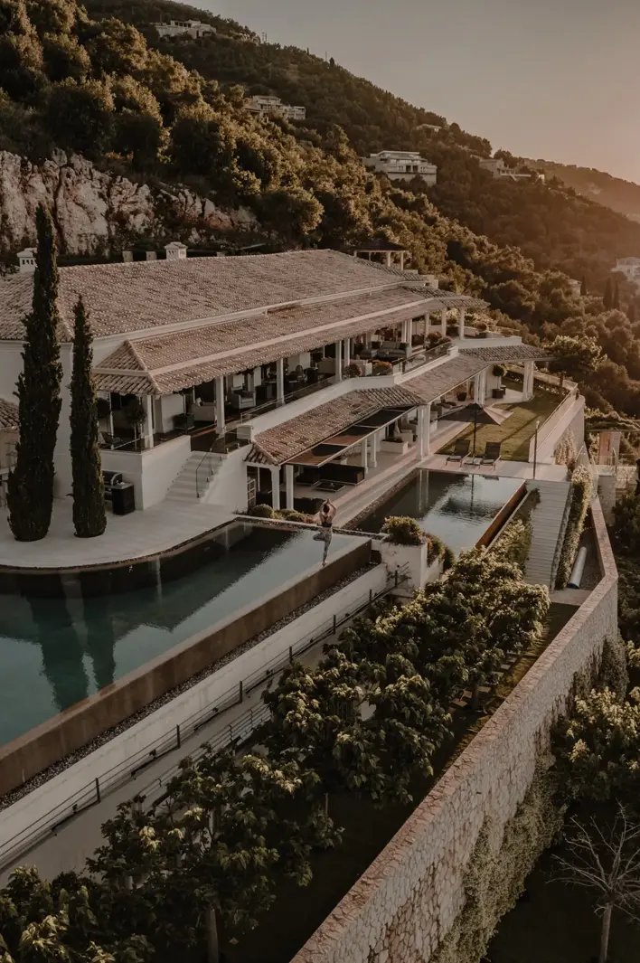 Aerial view of a luxury seaside villa built on a hillside, with terraces, pools, and a sunset over the ocean.