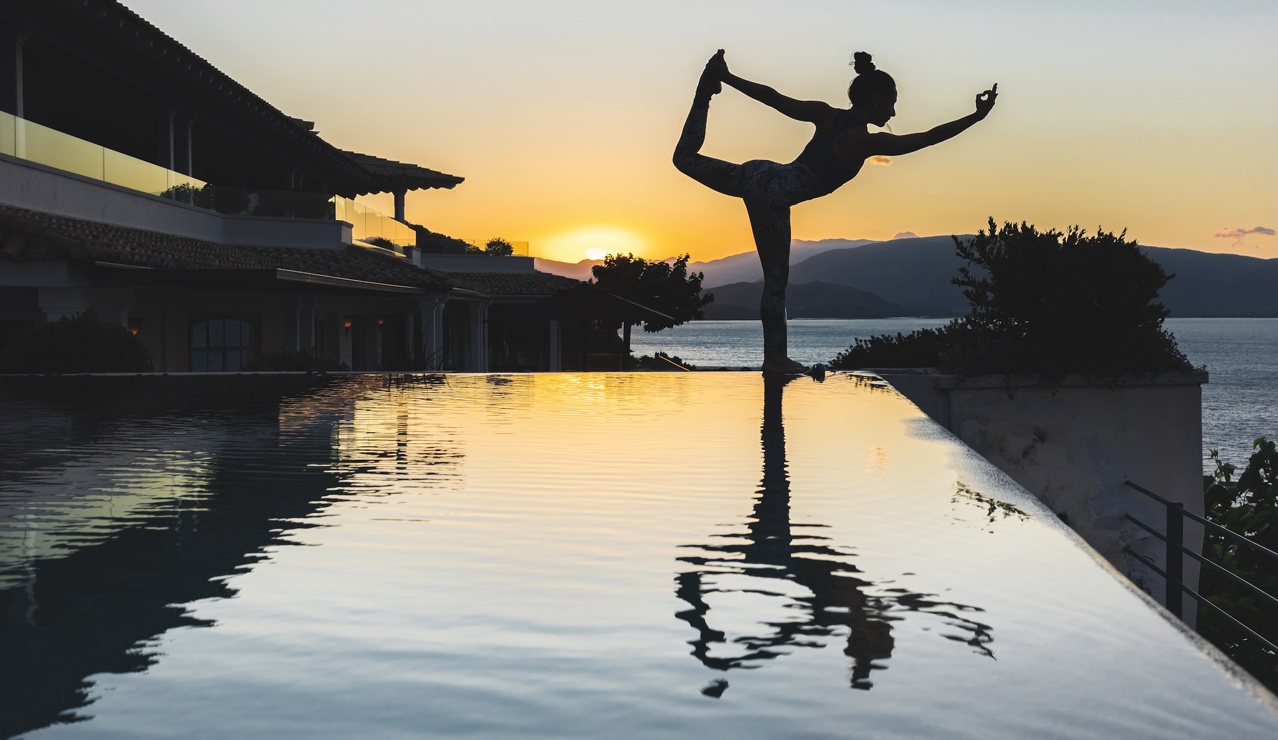 Silhouette of a woman practicing yoga by an infinity pool at sunset, with mountains and sea in the background.”