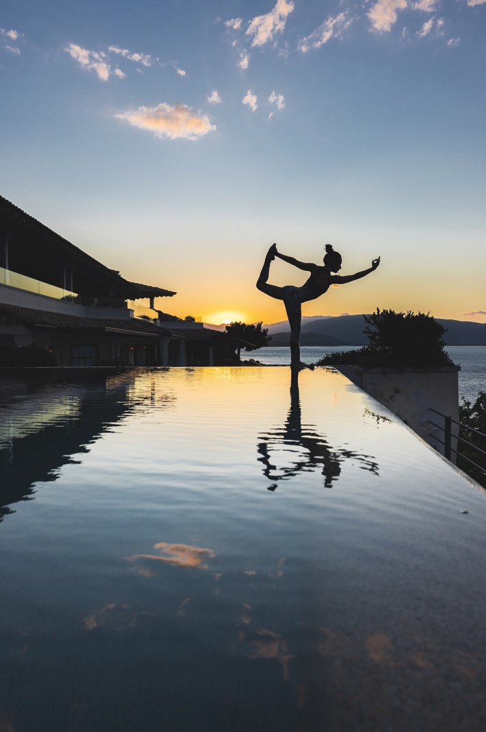 Silhouette of a woman practicing yoga by an infinity pool at sunset, with mountains and sea in the background.”