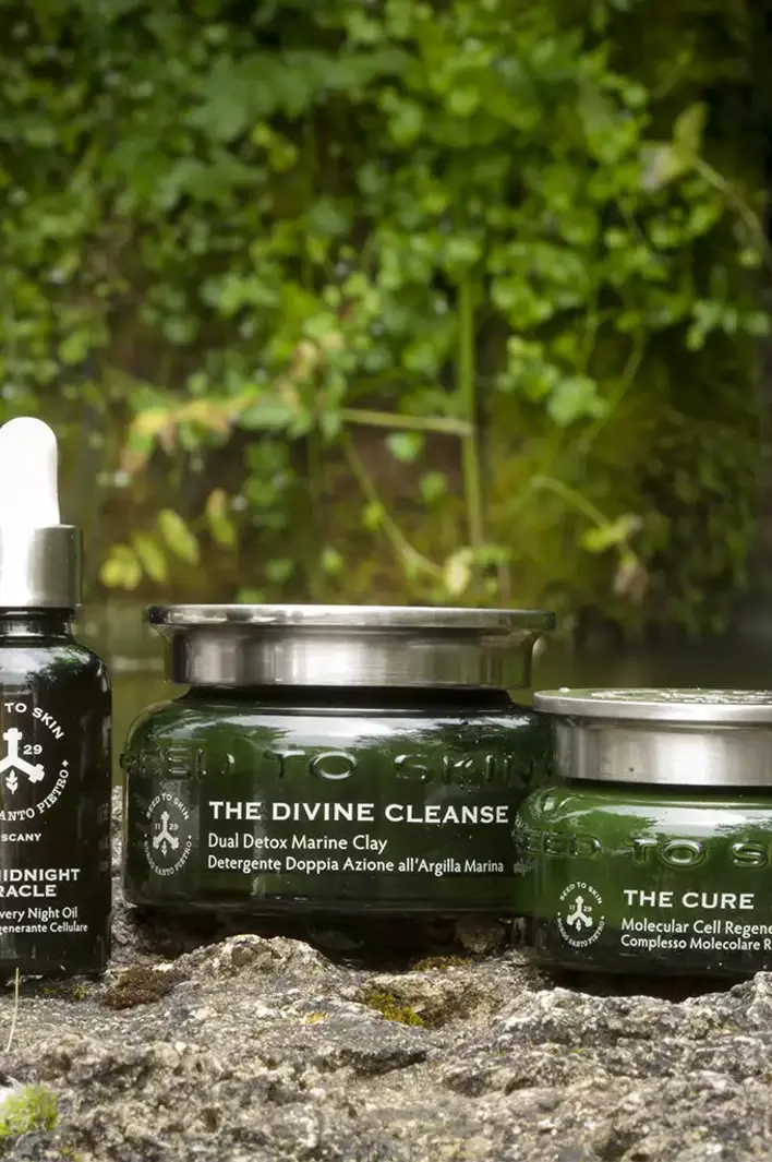 Seed to Skin Tuscany natural skincare collection including The Alchemist, The Midnight Miracle, The Divine Cleanse, and The Cure displayed outdoors by a waterfall.