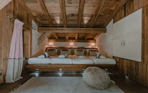 Cozy chalet children’s bedroom with wooden beams, soft lighting, plush toys, and multiple single beds in a warm alpine design.