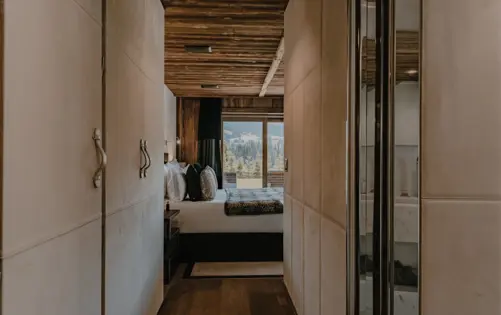 Modern luxury chalet bedroom with wooden ceiling, neutral tones, and mountain view through large window.