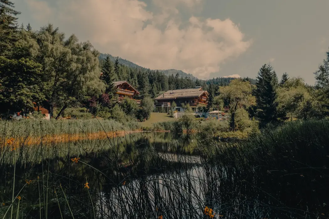 Lakeside alpine chalets surrounded by lush greenery and forested hills, reflecting in the calm water under a partly cloudy summer sky.