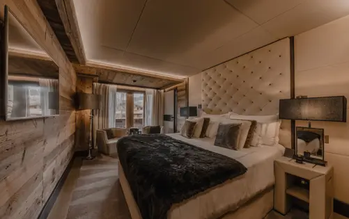 Elegant chalet-style bedroom featuring a large, plush bed with a tufted headboard, soft lighting, and cozy decor in warm neutral tones, with wooden walls and a view of the snowy mountains through glass doors.