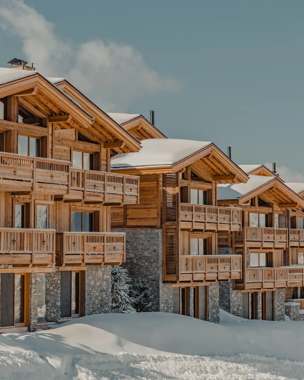 Row of luxury wooden chalets with stone facades covered in snow under a clear blue sky in the mountains.