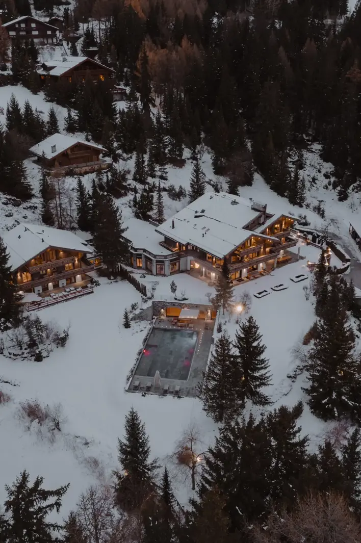 Aerial view of a luxury alpine chalet surrounded by snowy forest at dusk, with warm lights and a heated outdoor pool.”