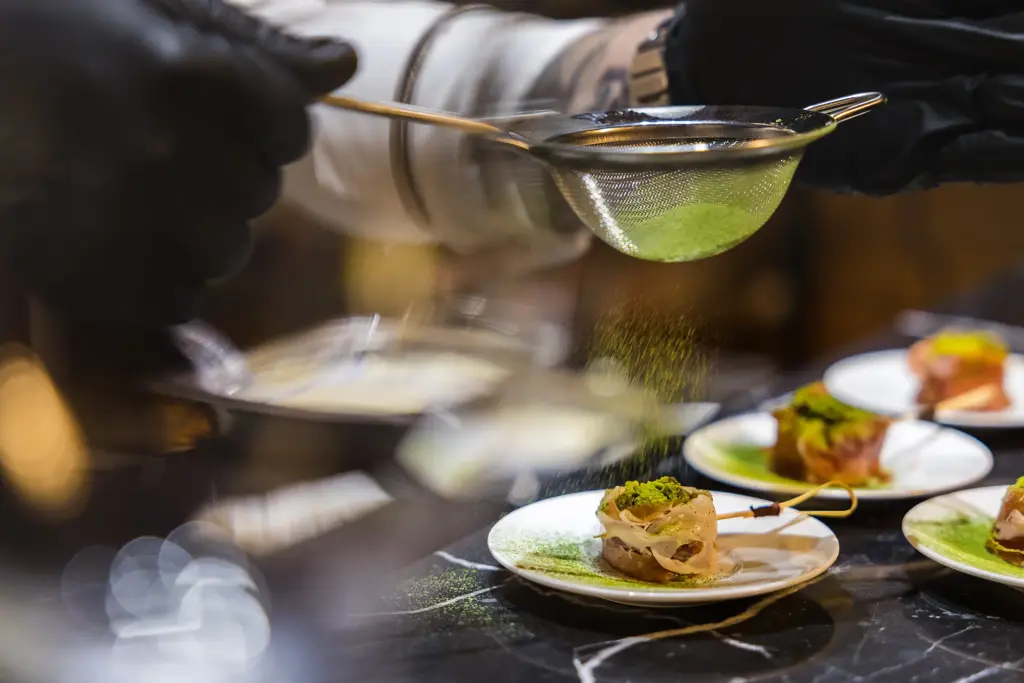 A chef wearing black gloves delicately adds the final touch to a gourmet dish, reflecting mastery, precision, and the artistry of fine dining.
