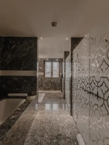 Luxurious marble bathroom featuring a deep bathtub, patterned glass shower partition, and a window providing natural light, creating an elegant and serene spa-like atmosphere.
