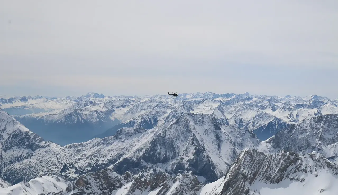 A helicopter flies above snow-covered alpine peaks on a clear winter day.