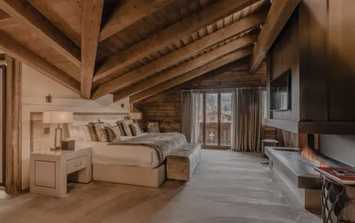 A luxurious chalet bedroom with a sloped wooden ceiling, a large bed dressed in soft linens and fur accents, and a cozy fireplace built into the wall. Natural light pours in through glass doors leading to a balcony with mountain views, creating a warm and inviting alpine atmosphere.