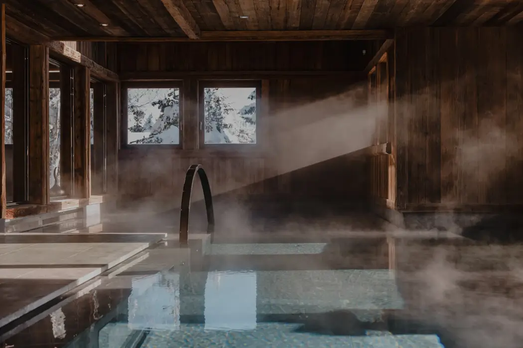 Luxury chalet indoor pool with wooden walls, sunlight streaming through windows, and steam rising for a cozy alpine spa atmosphere.
