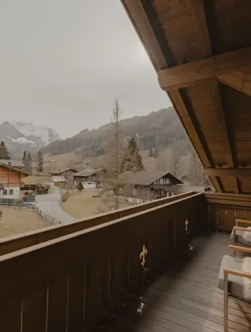 A cozy wooden balcony overlooking charming alpine chalets and misty mountain peaks. Soft fur throws on lounge chairs invite peaceful relaxation amidst nature.