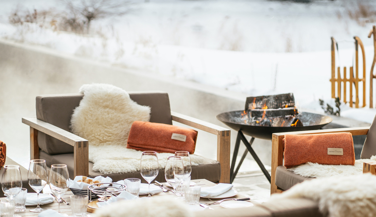 An outdoor lounge with fur-draped chairs and a firepit offers a cozy winter dining experience in a mountain setting.