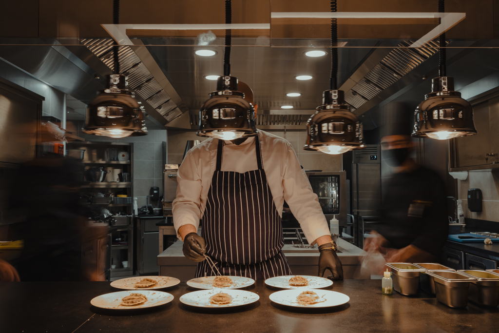 Chef in a professional kitchen plating gourmet dishes under warm pendant lights, with sous-chefs moving in the background.