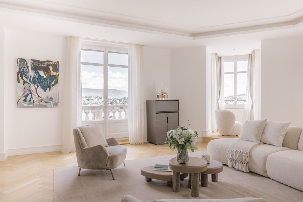 Bright modern living room with minimalist décor, light furniture, large windows overlooking a balcony, and soft natural lighting.