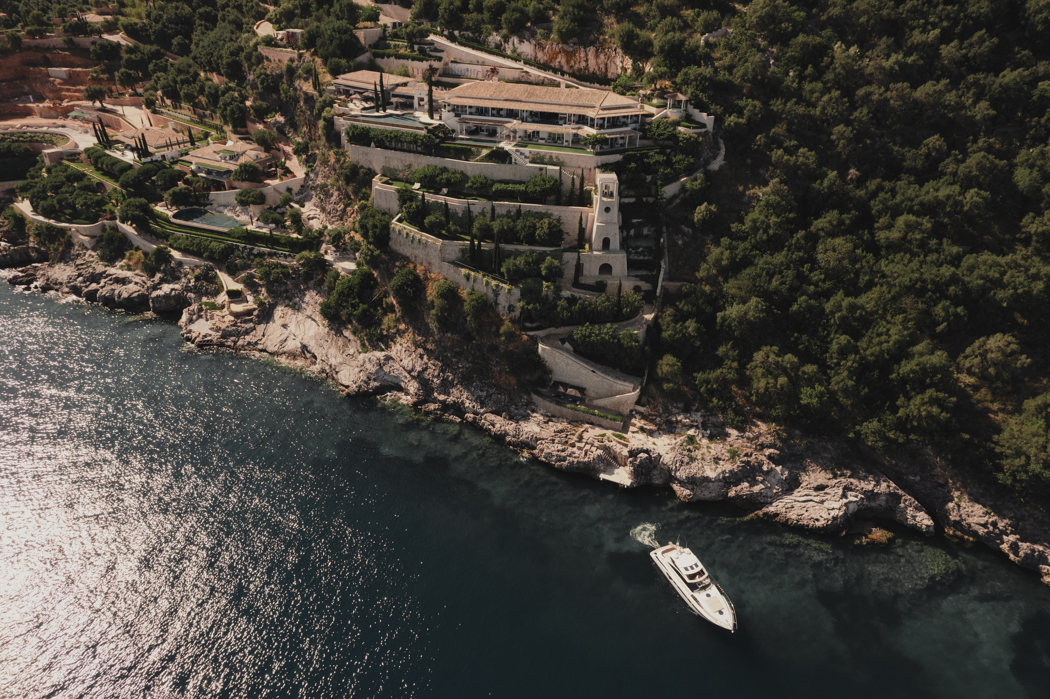 Aerial view of a luxury cliffside villa surrounded by lush greenery with a yacht sailing along the rocky coastline.